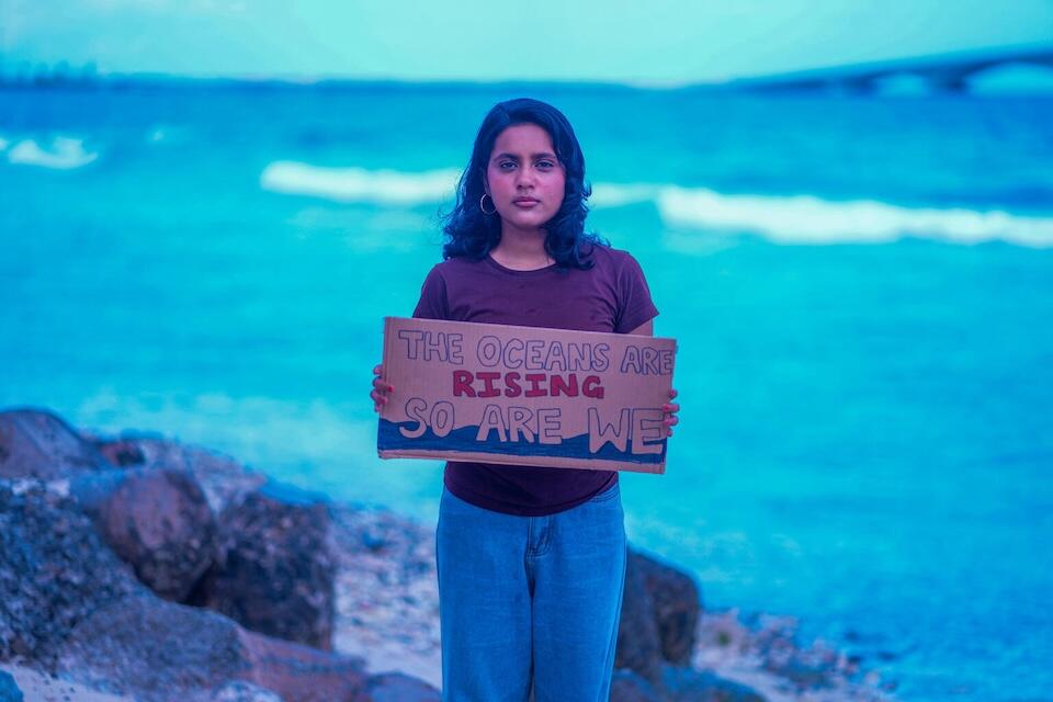 A young woman stands on a beach in Maldives holding a sign that reads "the oceans are rising, so are we."