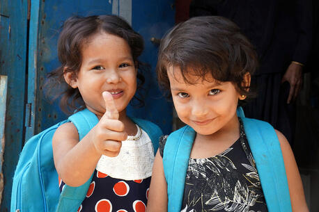 Two young smiling children wear blue UNICEF backpacks. One child gives a thumbs up.