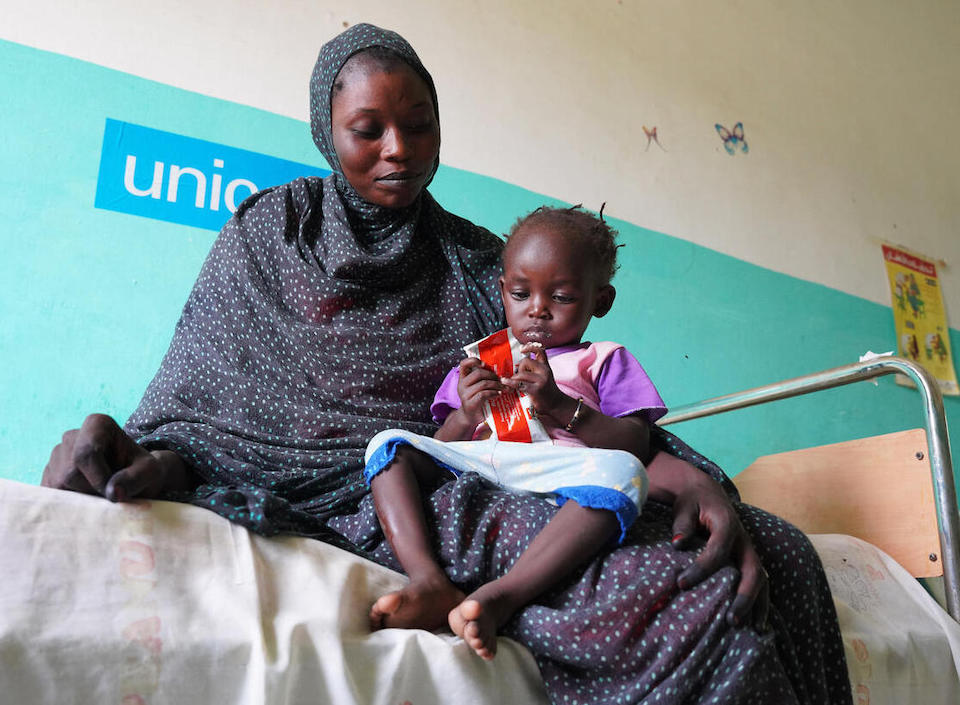 One-year-old Maub sits in her mother's lap holding a packet of Ready-to-Use Therapeutic Food (RUTF) at UNICEF-supported Abu Sunun Health Center, where she is being treated for malnutrition. 