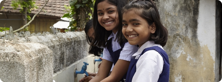 Three girls in school uniform overalls stand smiling at a stone outdoor water station, with their hands under its metal faucets.
