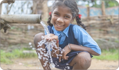 A girl squats in front of a water pipe opening, where she collects water in a container as it flows out.