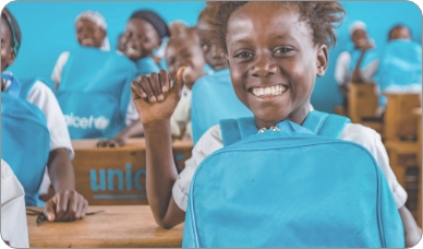 A classroom full of young female students displaying their blue UNICEF backpacks while smiling.