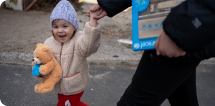 A smiling toddler holding a teddy bear walks holding her parent's hand.