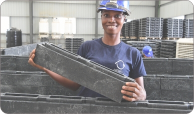 A smiling woman in a hard hat carries a small container in a large warehouse
