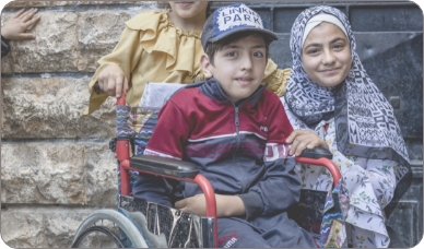 A boy sits in a wheelchair, while two girls stand smiling next to and behind him.
