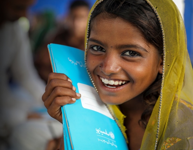 A female student in a headscarf smiles while holding a blue UNICEF workbook up to the side of her face.