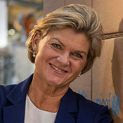 Bust portrait of Maria Ahlström-Bondestam, a woman with close cropped blonde hair, wearing a blue jacket over a white shirt, standing in a UNICEF warehouse.