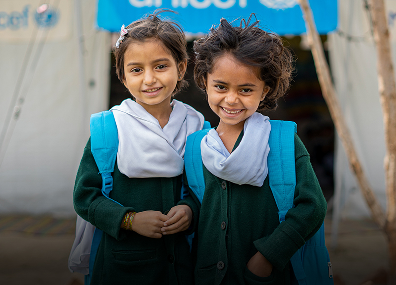 Two girls wearing blue UNICEF backpacks stand in front of a UNICEF tent.
