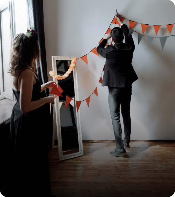 A man and a woman in fancy dress, decorate a room with Halloween colored banners.