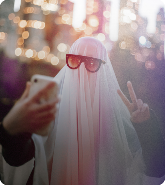 A trick-or-treater in a ghost bedsheet costume and sunglasses, gives a peace sign with their hand as they take a selfie.