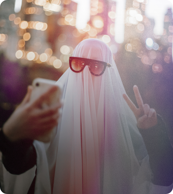 A trick-or-treater in a ghost bedsheet costume and sunglasses, gives a peace sign with their hand as they take a selfie.