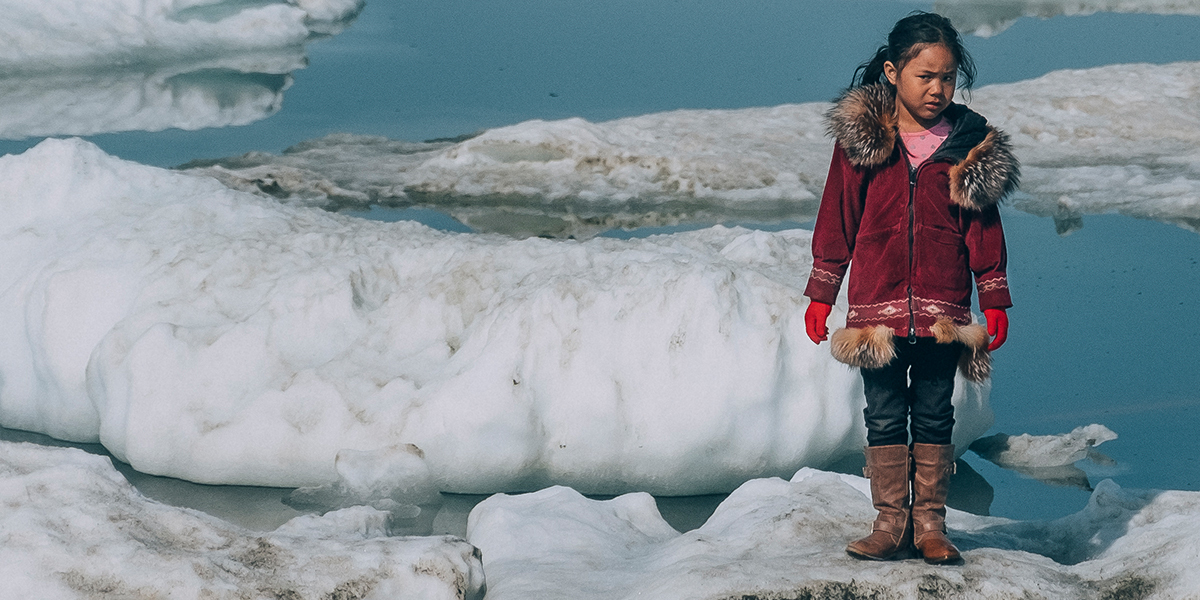 a girl wearing thick outerwear stands on an iceberg in ice-filled waters.