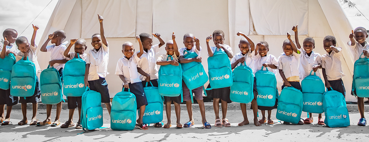 A large group of young students stand in front of a UNICEF tent, each holding up their blue UNICEF backpack.