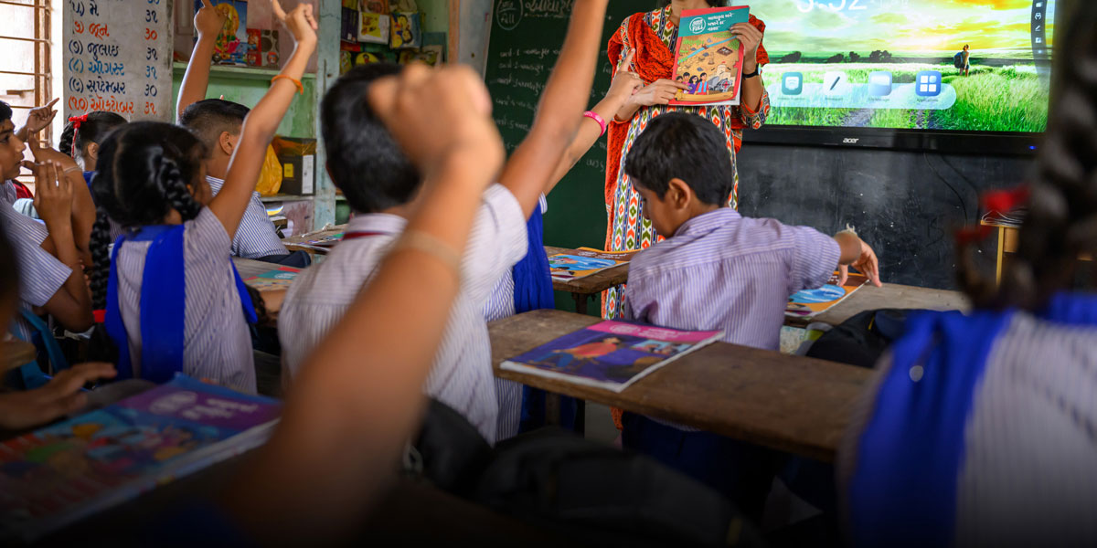 A classroom of children hold up their hands to participate in class.