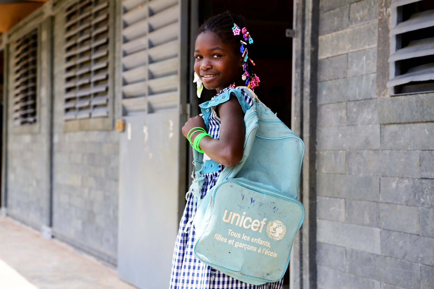 A schoolgirl walking into school, turns her head smiling back at the camera, while wearing a blue UNICEF backpack.