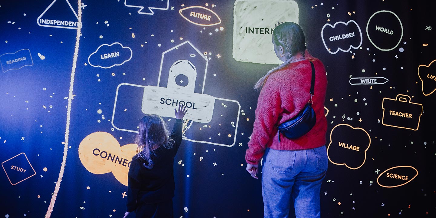 A woman and a young girl each put one hand on a wall covered in illuminated line drawings with labels like, School, internet, and learn.