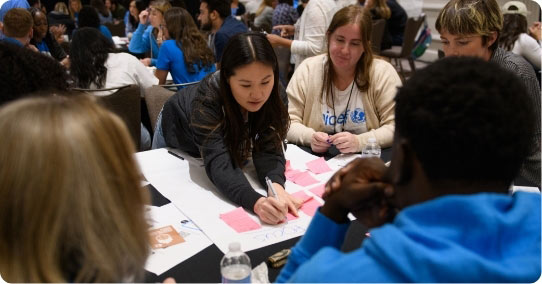 A group of students huddled over tables in a crowded room, working together over projects and post-it notes on the tables.