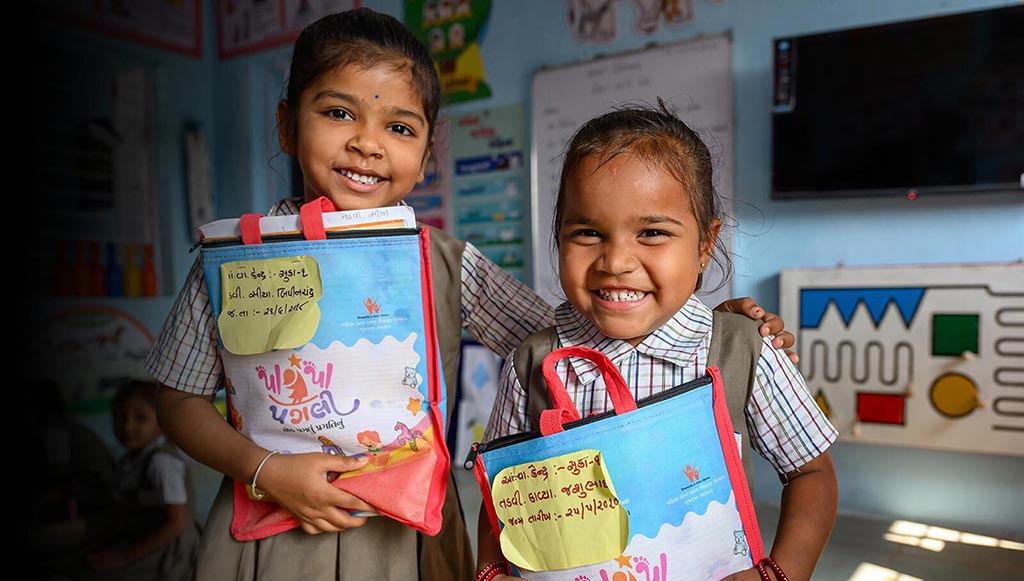 Two girls in a classroom smile while holding up UNICEF workbooks. The certification 'Great Place to Work, Certified, April 2024 to April 2025, USA' is pinned to the top right of the photo
