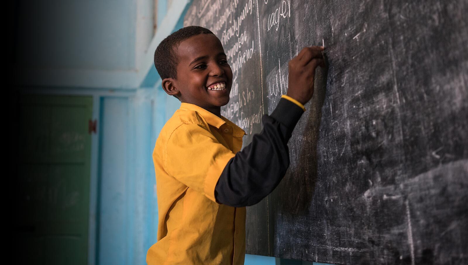 A smiling child in a yellow shirt writes on a blackboard at school. The certification 'Great Place to Work, Certified, April 2024 to April 2025, USA' is pinned to the top right of the photo