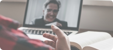 a hand in front of an open book and laptop that has a zoom window of a smiling man