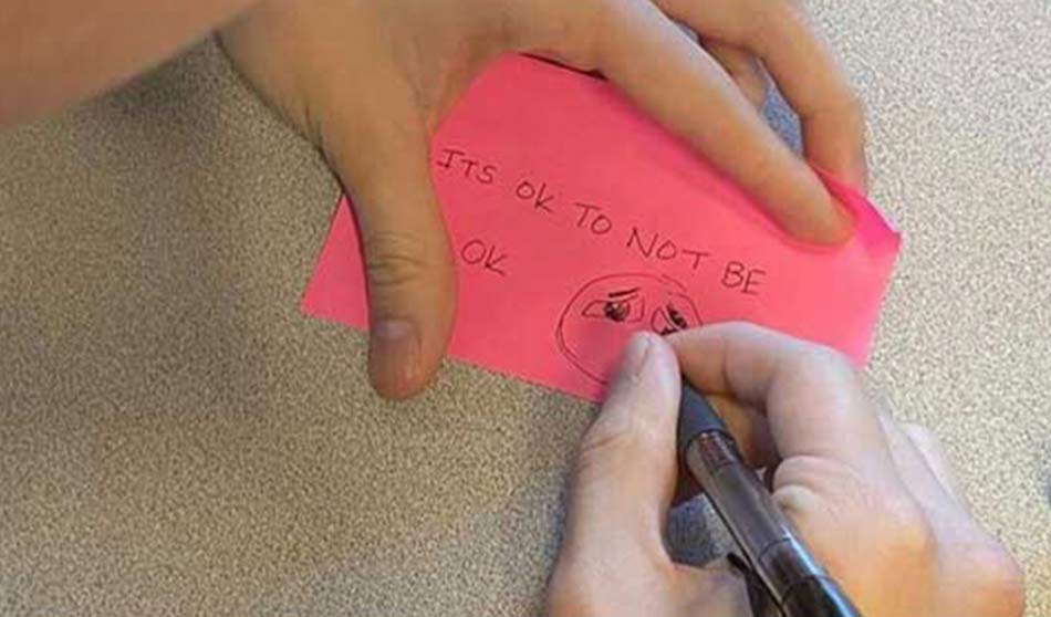 Hands write on a post-it note the words “It’s okay not to be okay” with a doodle of a sad face.