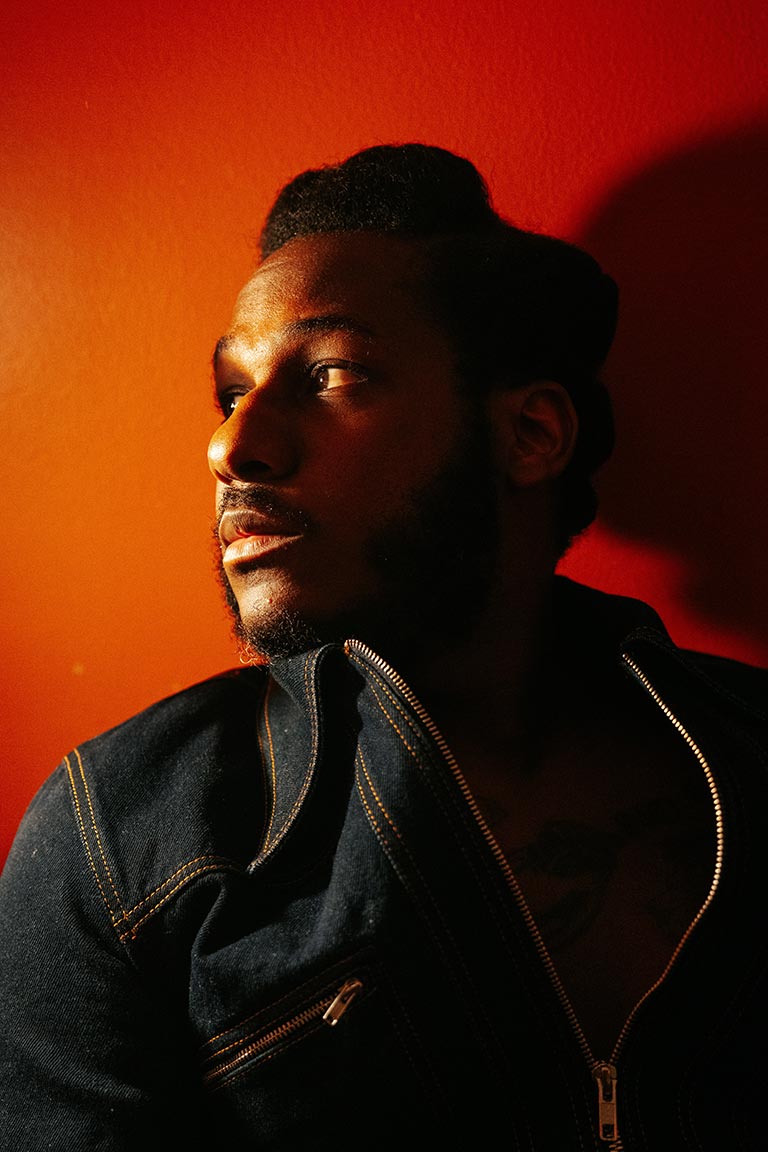 Headshot of Leon Bridges, in dramatic half-silhouette and open-front zip-up top, in front of a red-lit wall.