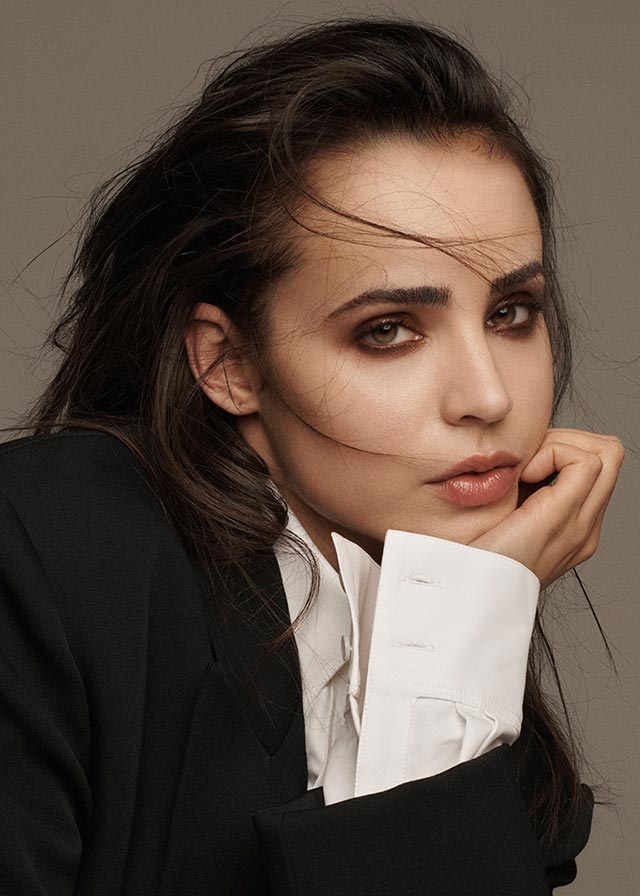 Profile photo of Sofia Carson in a suit, resting her head in her hand.