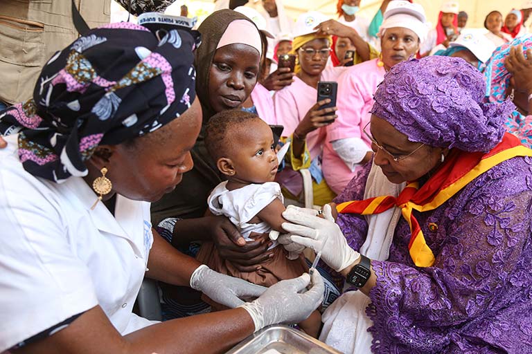 A woman wearing a head covering holds an infant while two female health workers inject a vaccine into the infant’s upper arm. Behind them several women stand watching and holding cellphones up to record.