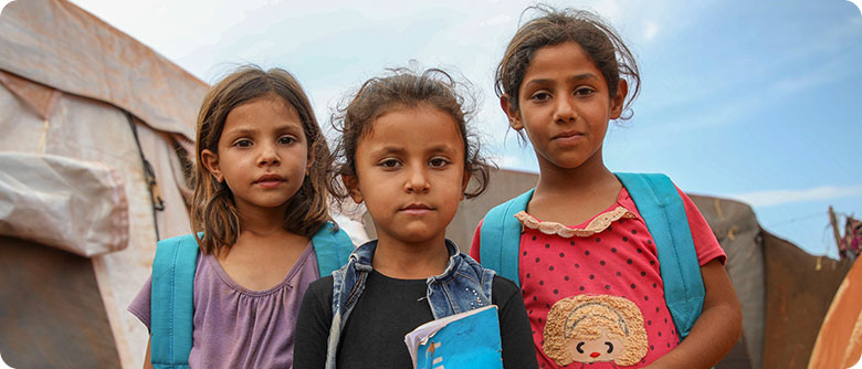 Three girls wearing blue UNICEF backpacks, stand in front of a UNICEF tent.