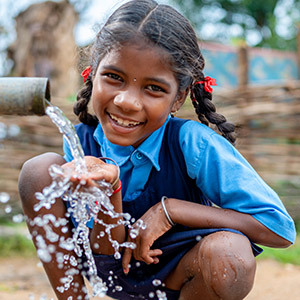 A girl in pigtails squats in front of a well pipe that dispenses water over her waiting hands.