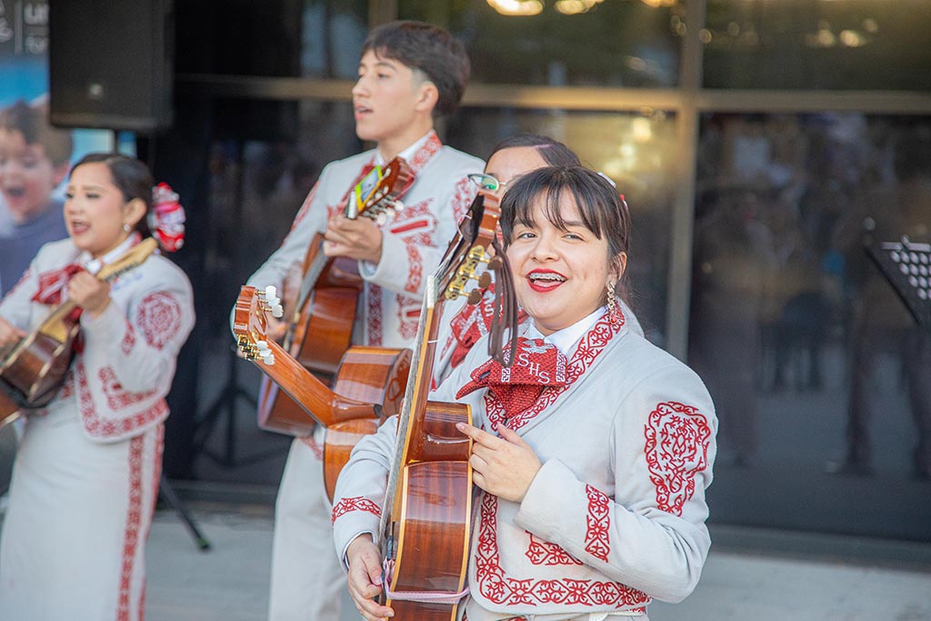 A mariachi band made up of three woman and a man play in front of a business building, a large speaker, and a UNICEF banner.