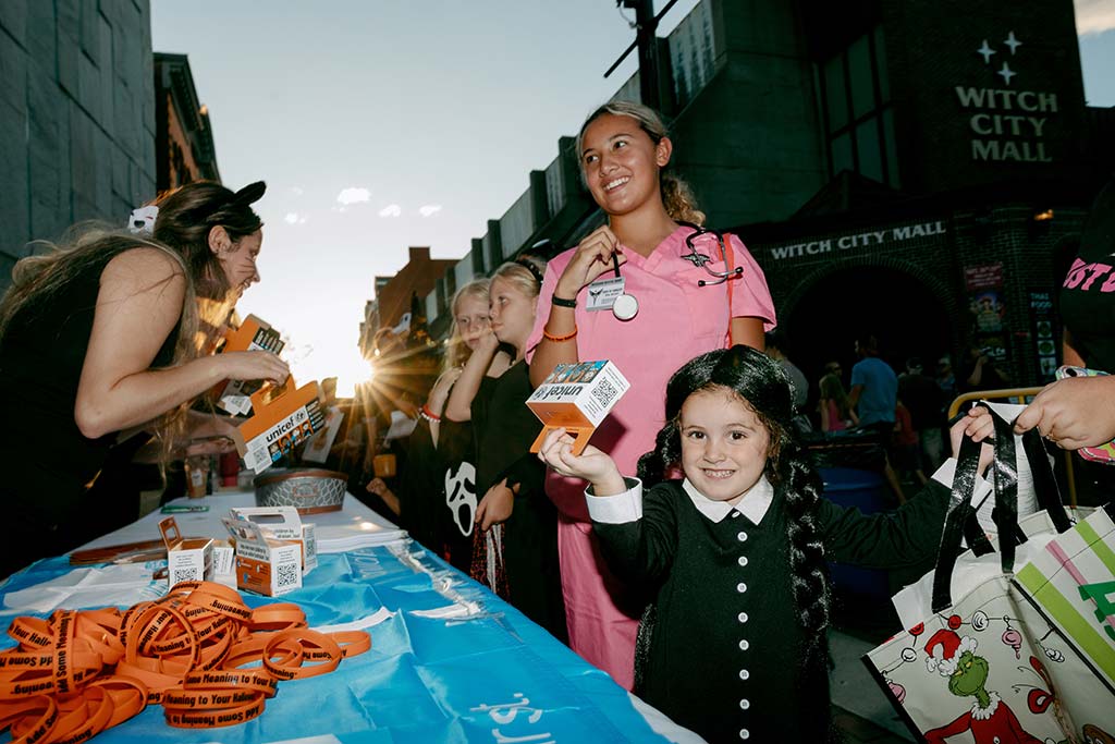 Children and adults in Halloween costumes collect trick-or-treat for UNICEF boxes at a UNICEF table in front of a building labeled "Witch City Hall"