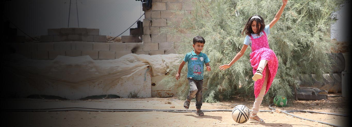 A girl in a pink dress plays football with a smaller boy, in front of a concrete wall and shrubbery outside