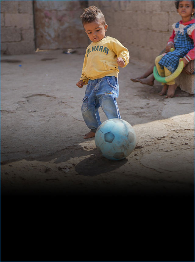 A toddler tries kicking a football outside in the dirt, while other children sit looking on.