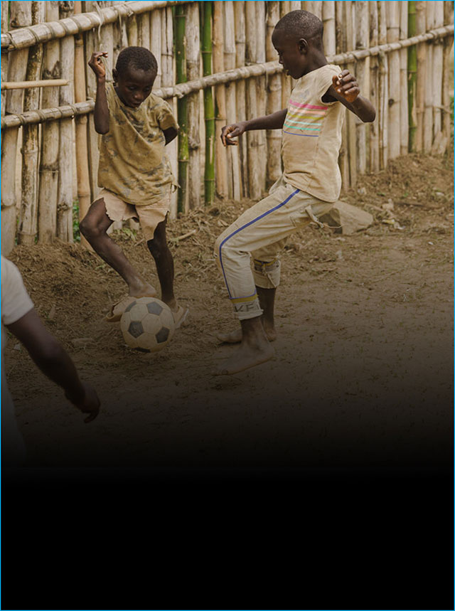 Two children play football in the dirt in front of a bamboo thatched wall.