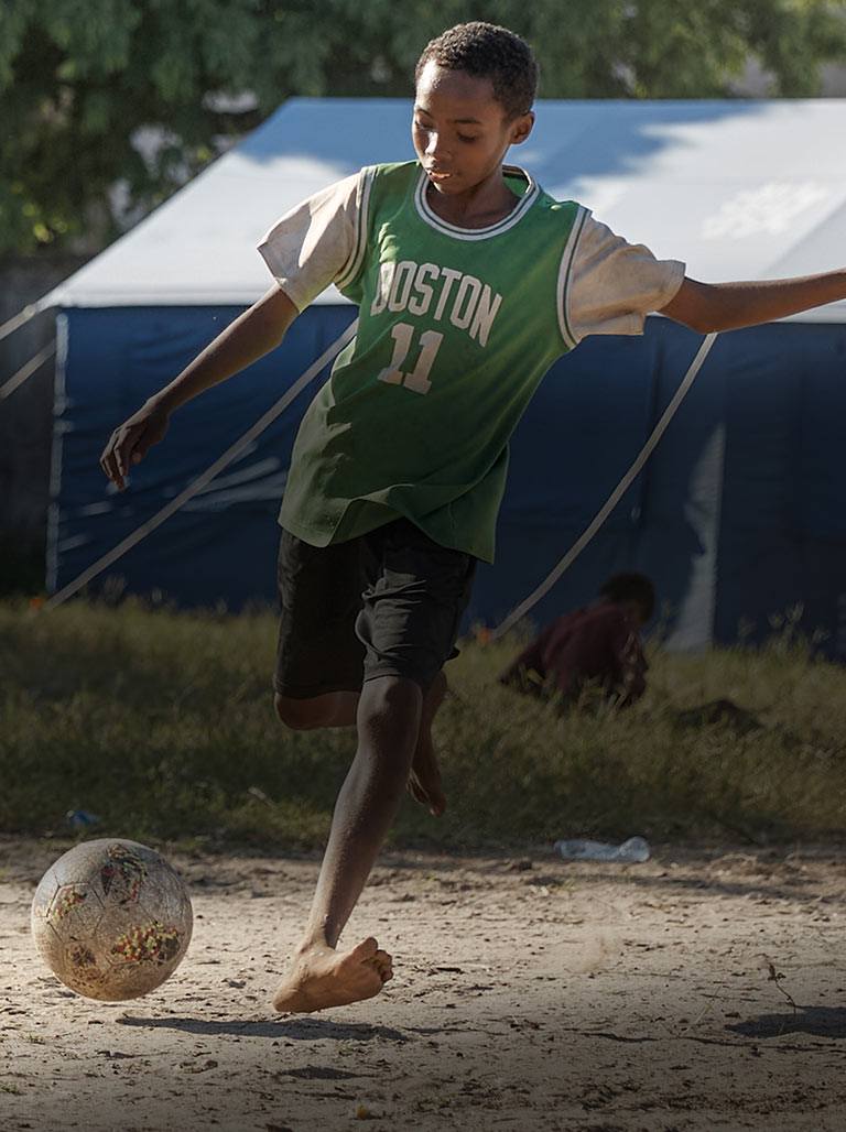 A boy in a green jersey plays football outside in front of a blue UNICEF tent