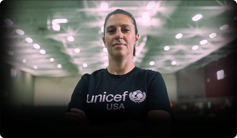 Carli Lloyd, a woman with her hair pulled back, stands in a UNICEF t-shirt in a warehouse.