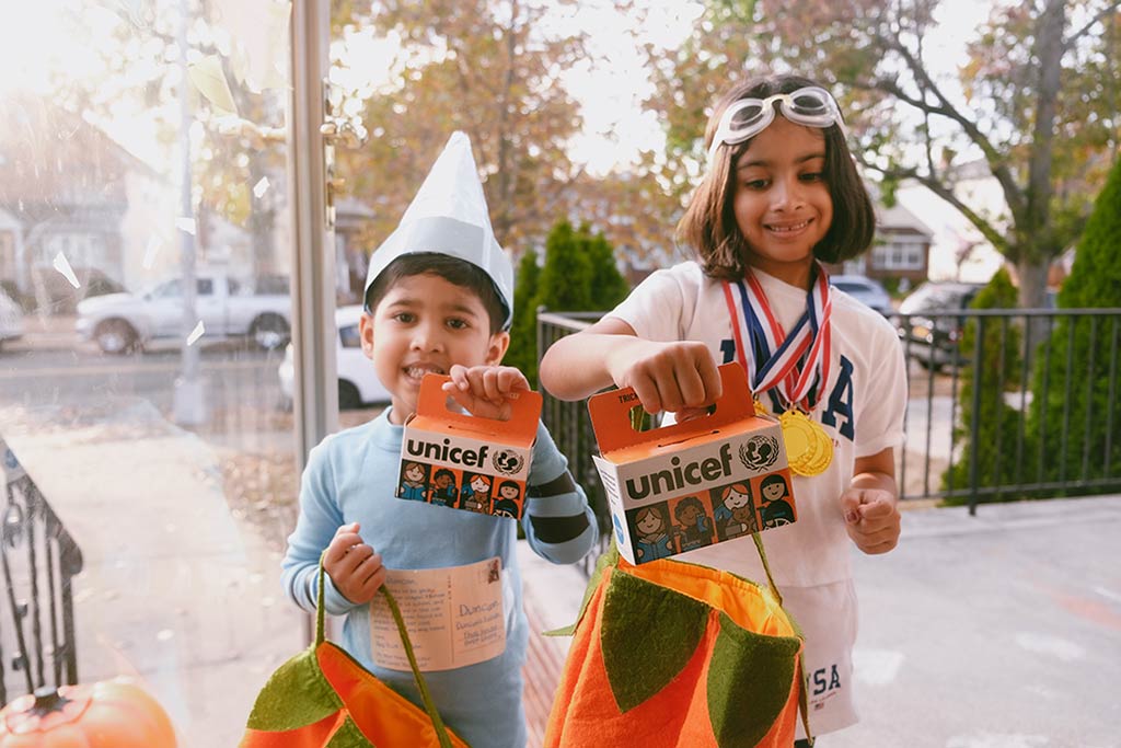 A boy in a paper hat, and a girl in swim goggles and Olympic medals over a USA tshirt, each hold up Trick-or-Treat for UNICEF boxes.