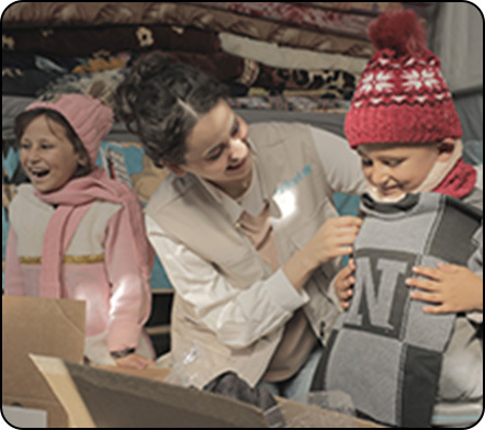 A UNICEF worker puts winter clothes from boxes, onto a smiling boy, while a girl laughs in her own winter clothing..