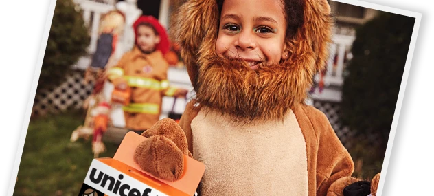 A child in a lion costume smiling and holding a Trick-or-Treat for UNICEF collection box