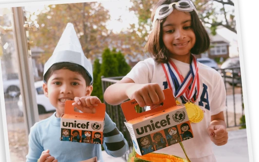 Two children in costume smiling and holding orange Trick-or-Treat for UNICEF collection boxes