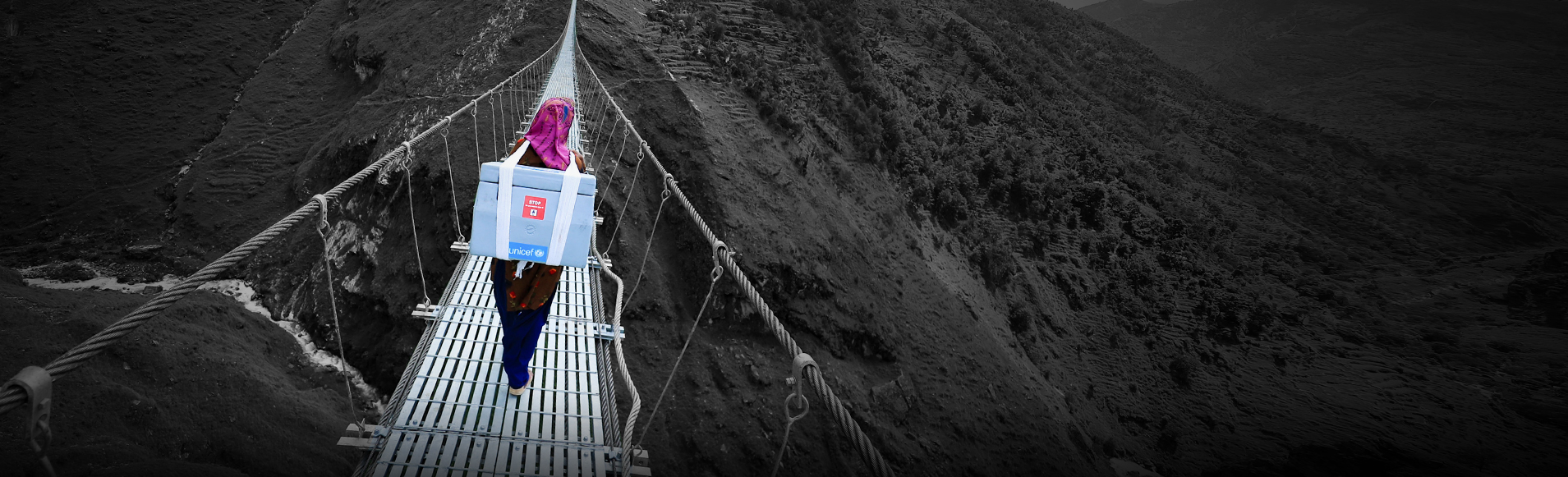 A woman carrying a UNICEF container on her back walks across a rope bridge between mountains