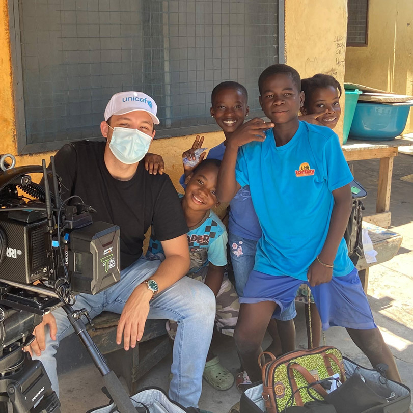 Director Ben Proudfoot, in a COVID mask, sits behind the camera in Accra, Ghana, surrounded by happy children, flashing peace signs.