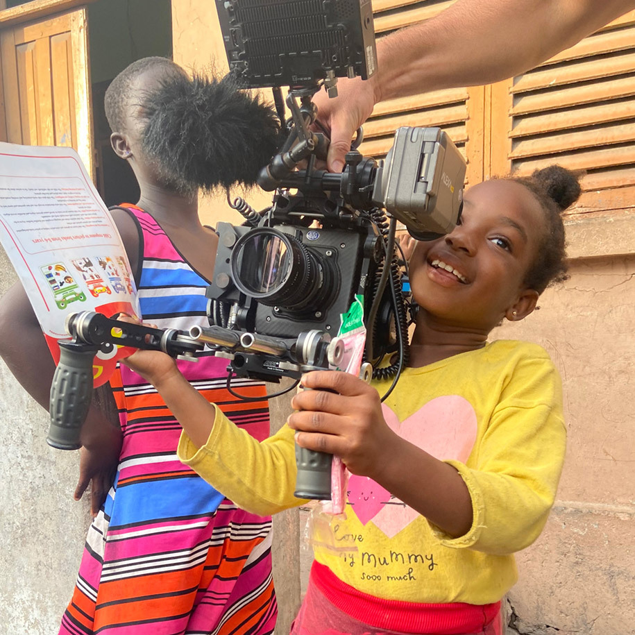 A young girl in Ghana, age five or six, pretends to be part of the film crew, and struggles to hold a large camera rig on her shoulder.