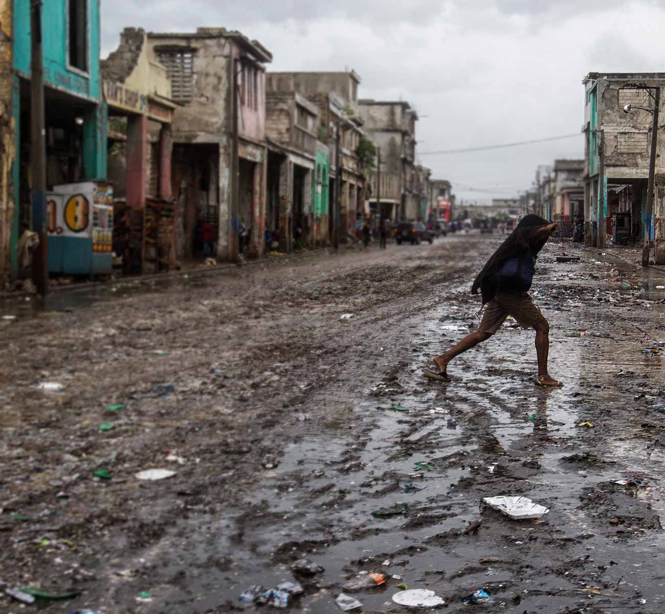 A Haitian boy in slippers crosses an unpaved road littered with mud and debris, and lined with ruined buildings - Donation form background
