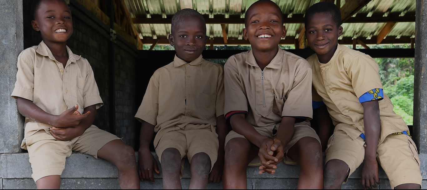 Four children sit smiling on the stoop of an open air building that is among trees