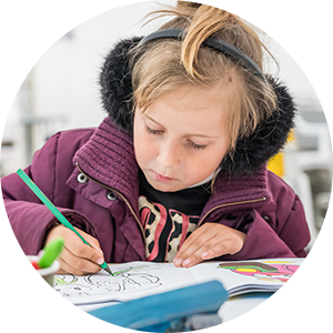 A child wearing earmuffs and a coat, sits at a school desk writing
