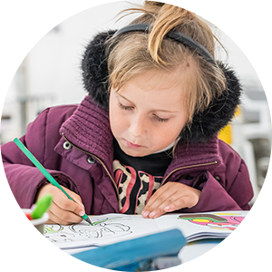 A child wearing earmuffs and a coat, sits at a school desk writing