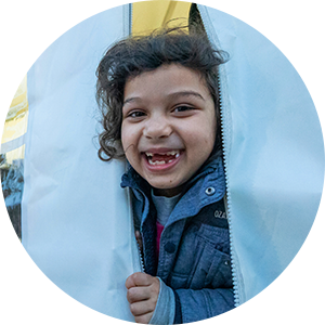A young child missing many teeth peeks out of the flaps of a tent, smiling and laughing
