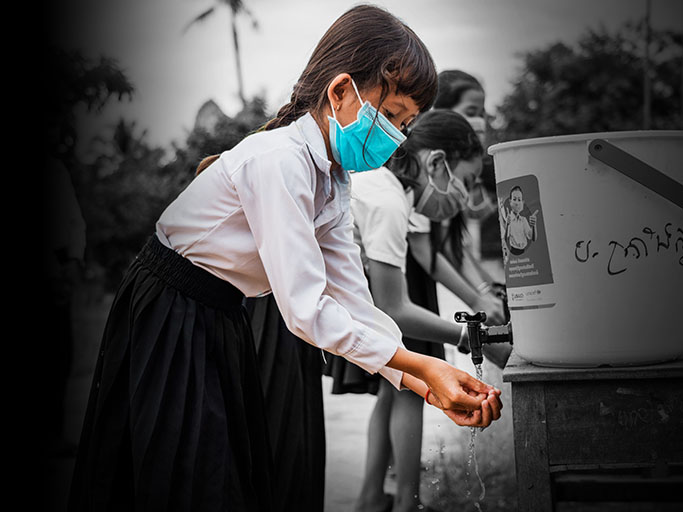 a girl washes her hands outside at a public water bucket dispenser with other children, all of whom wear face masks